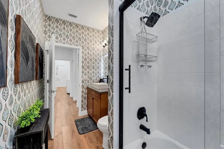 Bathroom with patterned wallpaper, wood-finish flooring, a vanity with a vessel sink, and a shower-over-tub with matte black fixtures