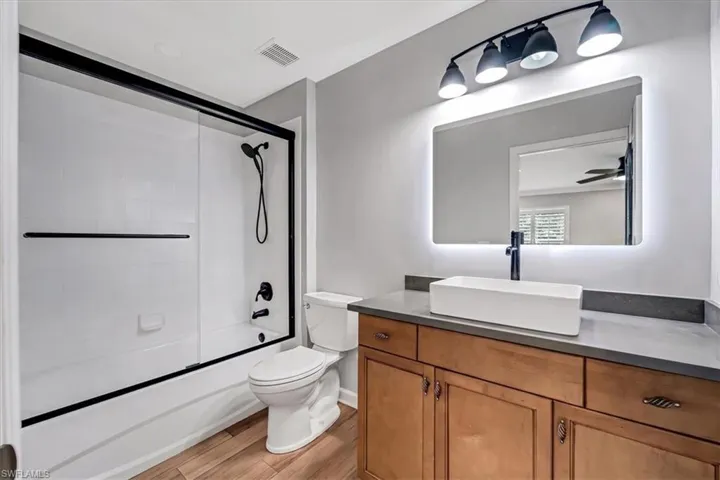 Wood-finish flooring, a contemporary vanity with a vessel sink, an illuminated mirror, and a tub with a sliding glass enclosure