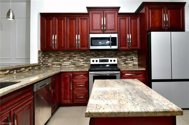 Kitchen with reddish brown cabinets, light stone counters, stainless steel appliances, and decorative light fixtures