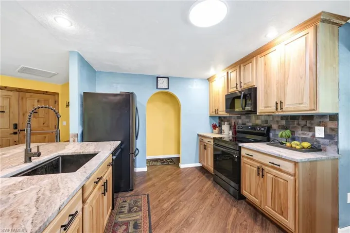 Kitchen with black appliances, arched walkways, light brown cabinetry, light stone counters, and dark wood-type flooring