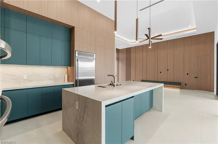Gourmet kitchen featuring a waterfall island with integrated sink, stainless steel refrigerator, and teal flat-panel cabinetry