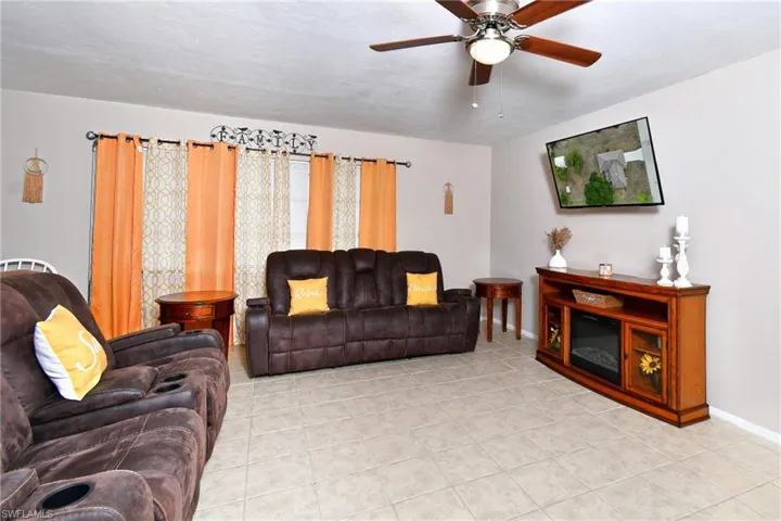 Bright living space featuring light-toned tile flooring, a ceiling fan with wood-finish blades, and a media console with an integrated electric fireplace