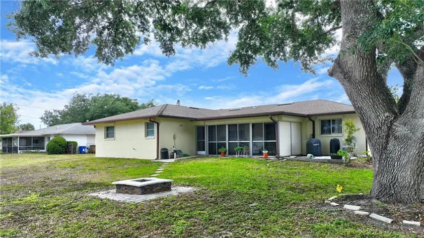 Rear exterior featuring a screened patio enclosure, a concrete patio area, a stone fire pit with paver surround, a mature tree, and a grass lawn