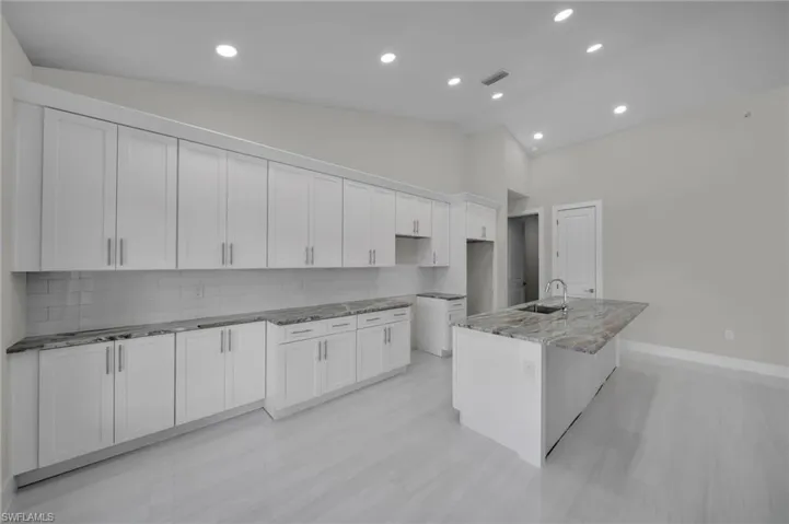 Kitchen featuring a sink, stone countertops, white cabinetry, backsplash, and a center island with sink