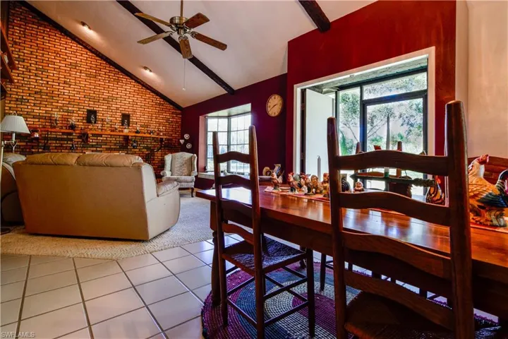 View Of The Great Room From The Dining Area Featuring Soaring Ceilings A Bricked Wall With Woodburning Fireplace And Hearth, Wood Beams, And Window Seat In The Bay Window Overlooking The Private Lake.