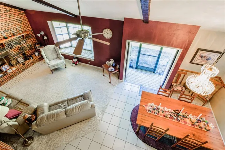 Birds Eye View Of The Great Room From The Balcony Overlooking The Dining And Great Room Areas Featuring A Ceiling Fan And Beautiful Glass Chandelier, Bricked Wall With Woodburning Fireplace And Hearth, Wood Beams, And Window Seat In The Bay Window Overlooking The Private Lake.