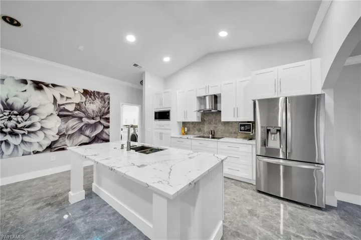 Kitchen with wall chimney exhaust hood, a kitchen island with sink, stainless steel appliances, white cabinetry, and light stone counters