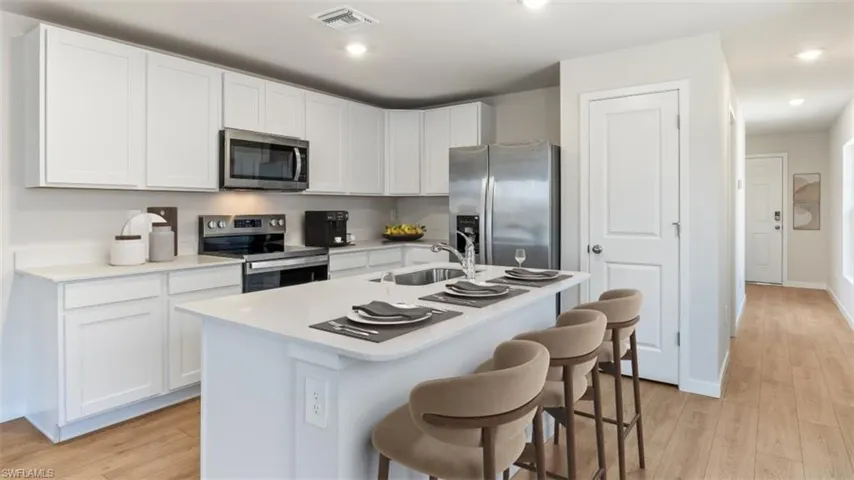 This image is virtually staged. Kitchen featuring stainless steel appliances, white cabinets, light wood-type flooring, a kitchen bar, and a center island with sink