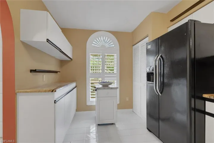 Kitchen featuring white flat-panel cabinetry, light-toned stone countertops, a tall arched window with fixed sunburst muntins and plantation shutters, louvered-door pantry storage, and tile flooring