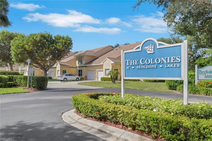 Community entrance featuring asphalt paving, landscaped hedging, mature trees, and multi-unit residential buildings with attached garages