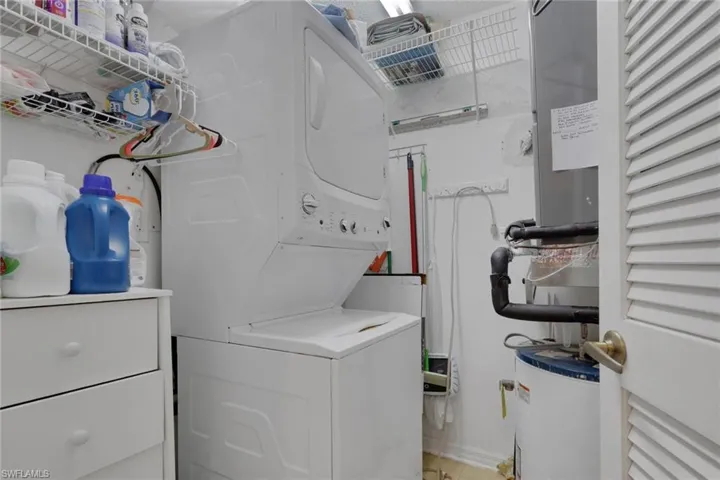 Utility closet featuring stacked laundry appliances, an electric water heater, built-in wire shelving, and louvered door access