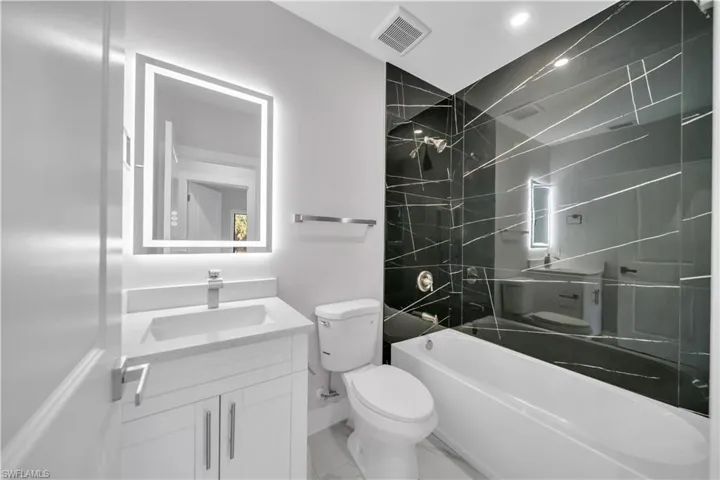 Contemporary bathroom featuring a black accent wall with linear accents, a white bathtub, a vanity with a white countertop and integrated sink, a backlit mirror, and a modern toilet