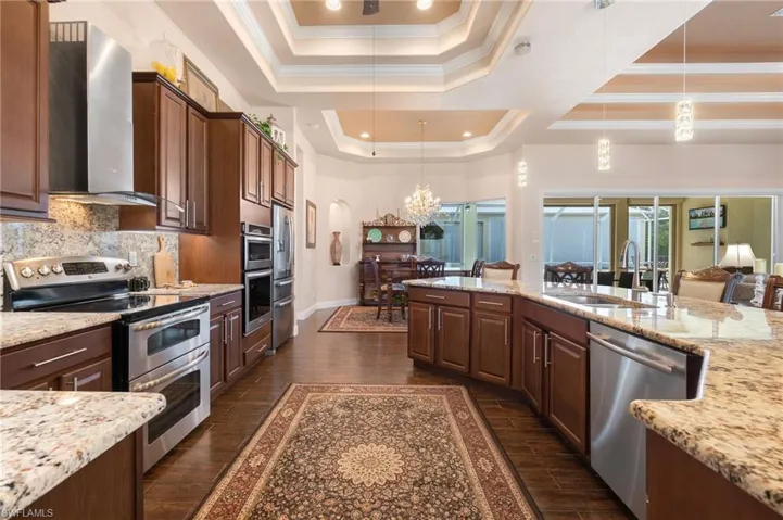 Kitchen with dark hardwood / wood-style floors, stainless steel appliances, hanging light fixtures, wall chimney exhaust hood, and a raised ceiling