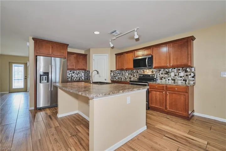 Kitchen featuring stainless steel appliances, light wood-type flooring, rail lighting, a center island with sink, and backsplash