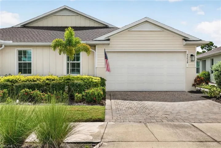 Light-colored exterior with a shingled roof, paver driveway, white garage door, and established landscaping