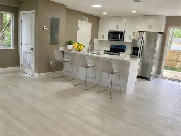 Open-concept kitchen featuring white shaker cabinetry, stainless steel appliances, a center island with bar seating, light wood-finish flooring, and a sliding glass door to the exterior