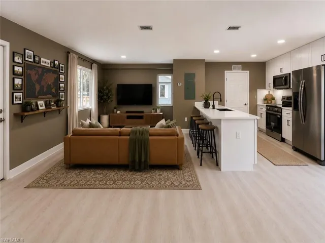 Open-concept living space featuring light wood-finish flooring, recessed lighting, a white kitchen island with seating, white shaker cabinetry, and stainless steel appliances