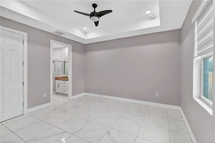 Unfurnished bedroom featuring ceiling fan, ensuite bath, a raised ceiling, light marble finish floors, and recessed lighting