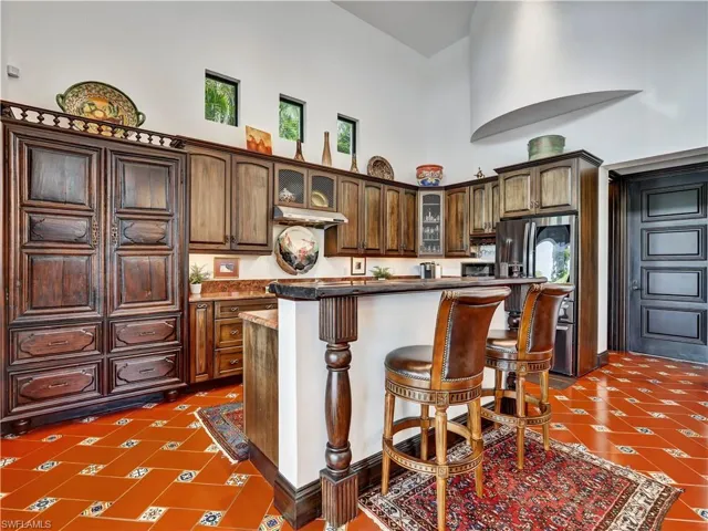 Kitchen featuring terracotta tile flooring with decorative accents, custom wood cabinetry, a built-in refrigerator, a breakfast bar with a decorative newel post, and high ceilings