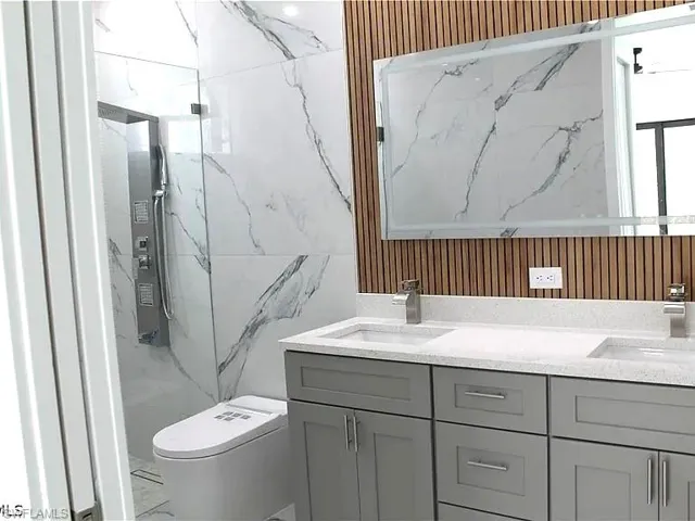 Bathroom featuring a dual vanity with light gray cabinetry and white speckled countertops, a large illuminated mirror, a wood-slat accent wall, and a walk-in shower with large format marble-look tile