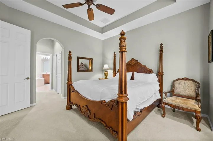 Bedroom with light carpet, a raised ceiling, arched walkways, a ceiling fan, and ensuite bath