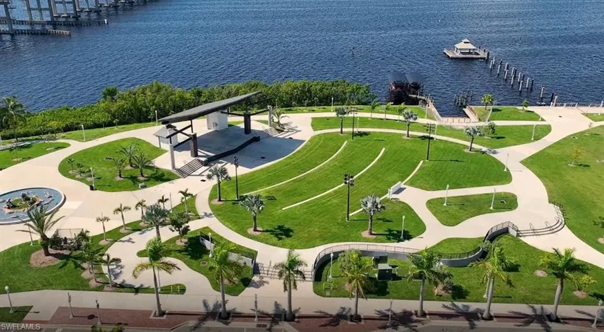 Waterfront park featuring a performance stage, paved walking paths, grassy terraced seating, and mature palm trees