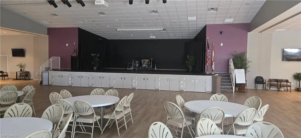 Spacious event hall featuring a raised stage with black backdrop, wood-finish flooring, acoustic ceiling tiles, and track lighting
