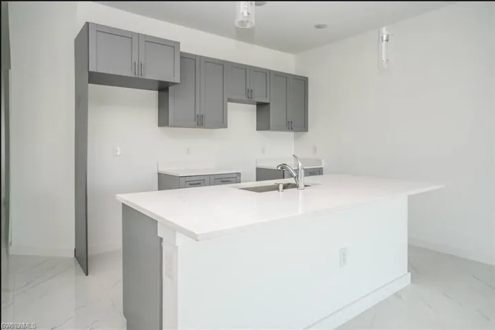 Kitchen featuring light marble finish floors, decorative light fixtures, gray cabinets, an island with sink, and light stone counters