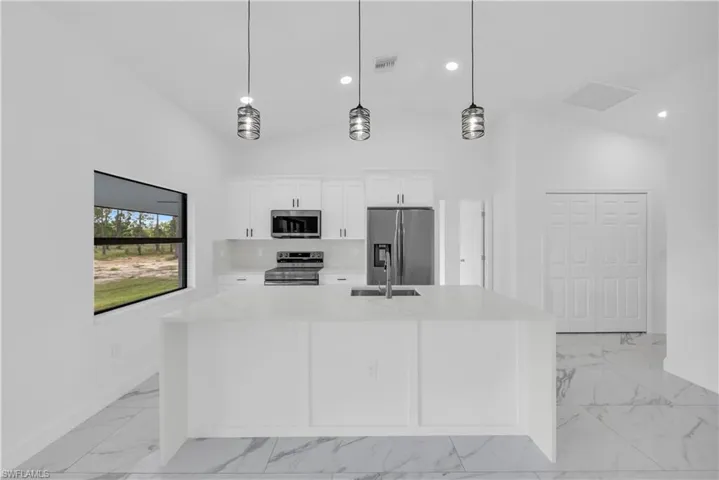 Kitchen with appliances with stainless steel finishes, light stone countertops, white cabinets, pendant lighting, and an island with sink