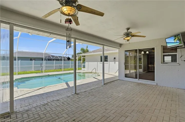 Covered patio featuring a herringbone-patterned paver floor, dual ceiling fans, sliding glass doors, and a screen-enclosed pool area with a white vinyl perimeter fence