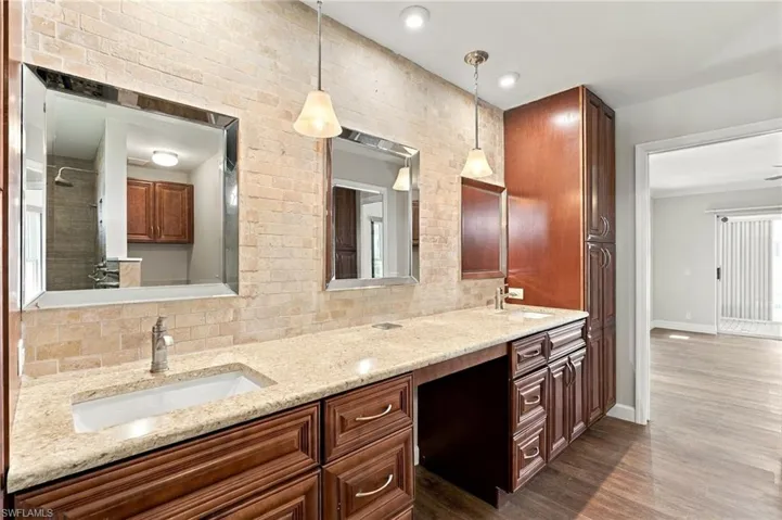 Dual vanity featuring stone-finish countertops, custom stained-wood cabinetry, brick-textured backsplash, framed mirrors, and pendant lighting