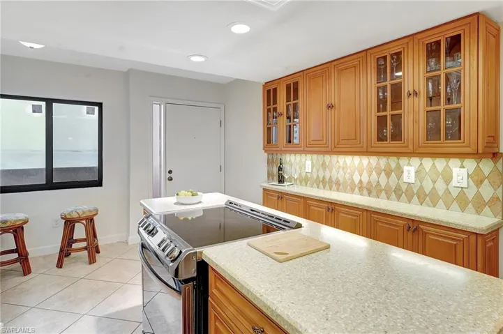 Kitchen with stainless steel electric stove, brown cabinetry, backsplash, light tile patterned floors, and light stone counters