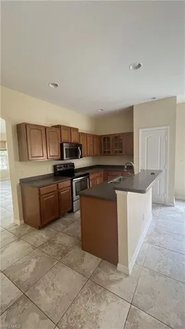 Kitchen with glass insert cabinets, stainless steel appliances, brown cabinetry, dark countertops, and a kitchen island with sink