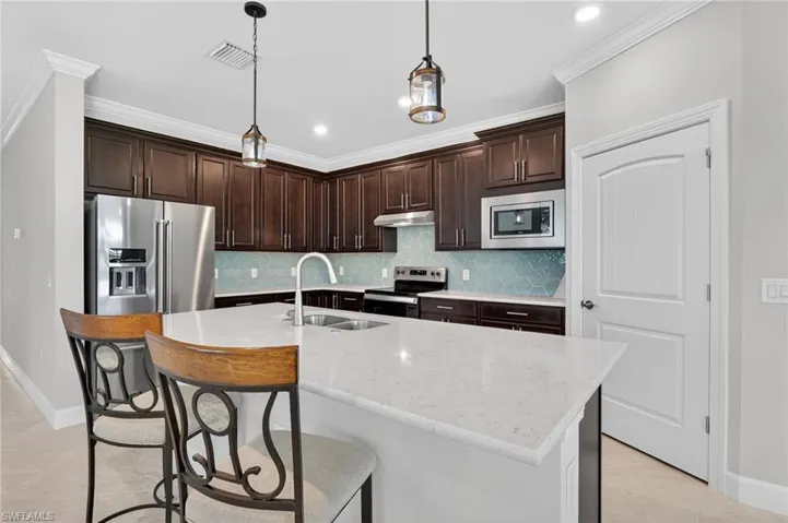 Kitchen featuring dark brown cabinetry, stainless steel appliances, a center island with sink, light stone counters, and pendant lighting