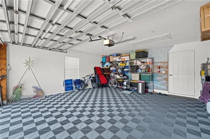 Spacious garage featuring checkerboard-patterned flooring, exposed ceiling beams, white textured walls, a single window with horizontal blinds, and a white paneled entry door