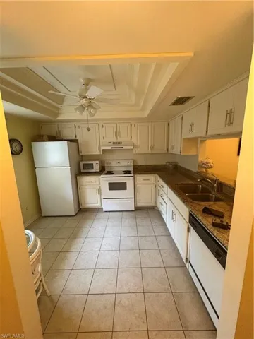 Kitchen featuring white cabinetry, granite-style countertops, stainless steel double sink, white appliances, and a tray ceiling with a ceiling fan
