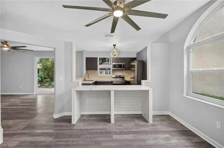 Wood-finish flooring, contemporary ceiling fan, kitchen featuring dark cabinetry and stainless steel appliances, breakfast bar with subway tile accents, and an arched window