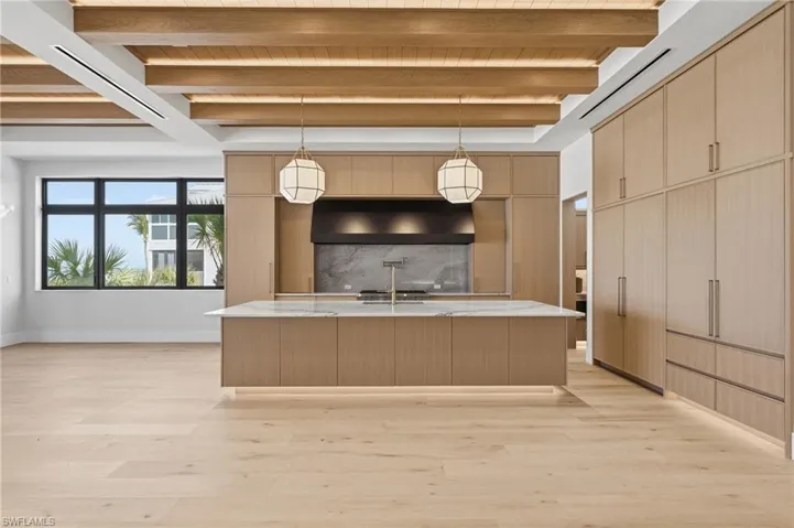 Gourmet kitchen featuring a large central island with a waterfall countertop, custom wood-finish cabinetry, and light wood-finish flooring - Virtually Edited Image