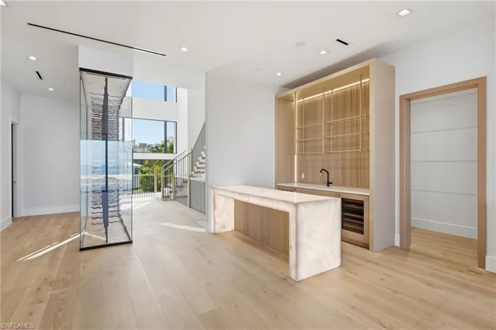 Open concept living area with wood-finish flooring, a built-in wet bar featuring a beverage cooler and illuminated shelving, a translucent stone countertop, and a glass-enclosed wine display
