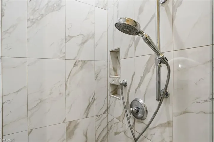 Shower enclosure featuring large format white tiled walls with grey veining, a chrome shower head with a flexible hose, and two recessed wall niches