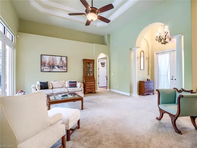 Living room with ceiling fan with notable chandelier, a tray ceiling, light colored carpet, and ornate columns