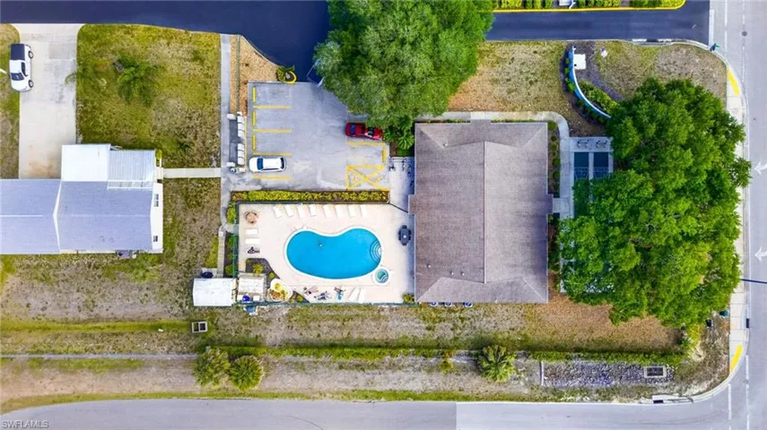 Aerial view of the property showing a large swimming pool with surrounding lounge chairs, a parking area, and a building with a dark roof