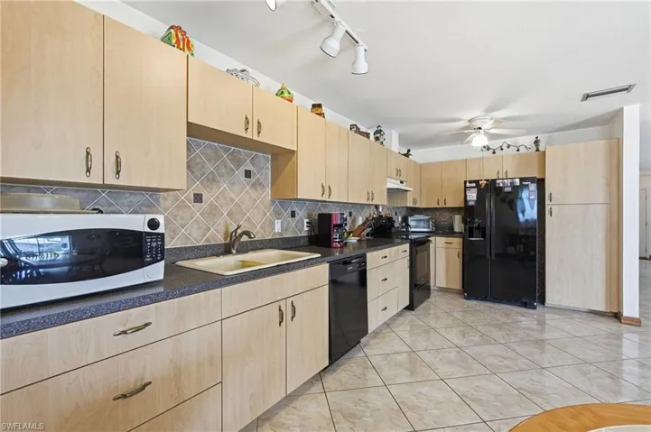 Spacious kitchen featuring extensive light wood cabinetry, dark countertops, a large black refrigerator, a tile backsplash with decorative accents, and neutral floor tiling
