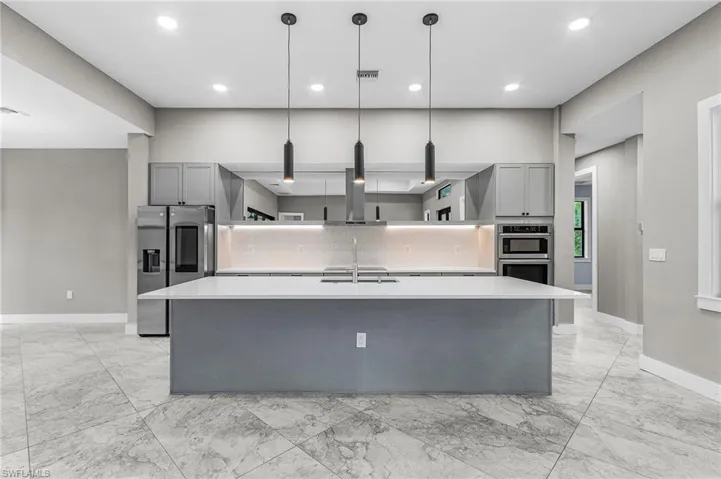 Kitchen with gray cabinetry, a large island with sink, decorative light fixtures, appliances with stainless steel finishes, and light marble finish flooring