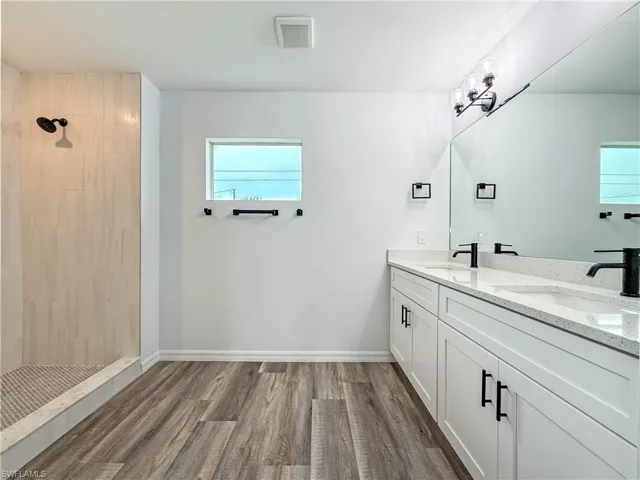 Full bath featuring tiled shower, double vanity, and dark wood-type flooring