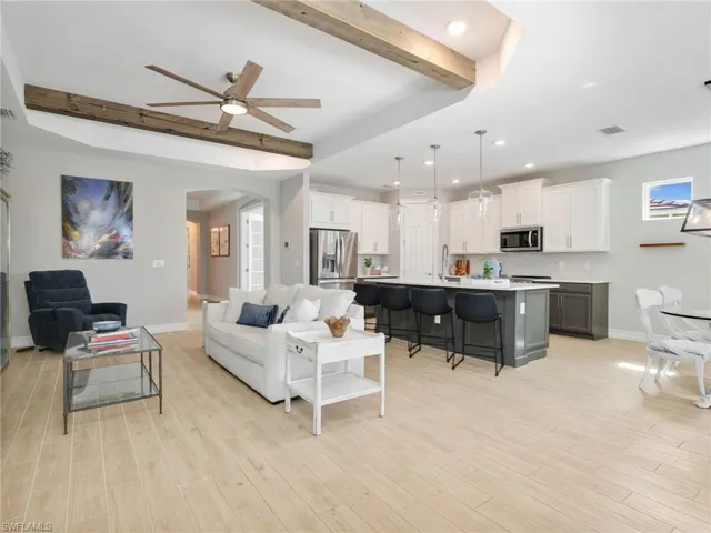 Open-concept living space featuring wood-finish flooring, a kitchen island with seating, pendant lighting, white shaker cabinetry, and exposed wood beams