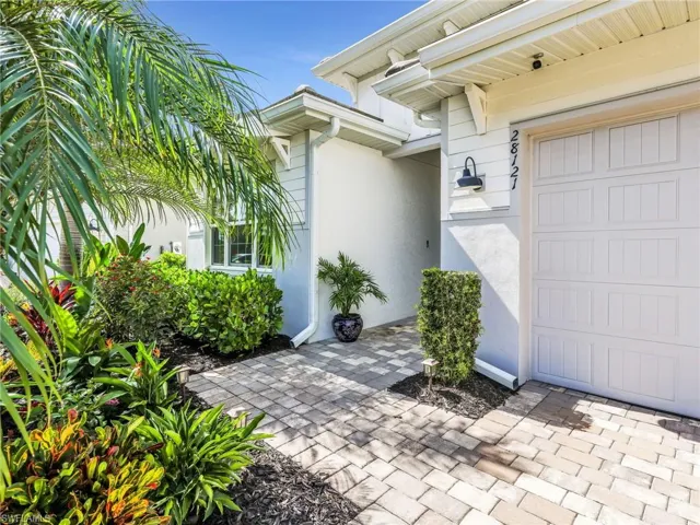 Front exterior showcasing a paver walkway, lush tropical landscaping, a white paneled garage door, and a wall-mounted exterior light fixture