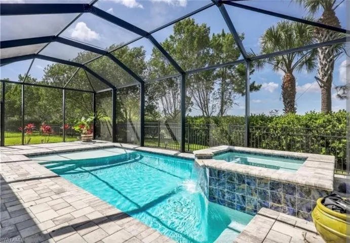 Screened enclosure featuring a swimming pool, integrated spa with waterfall, paver decking, and mature tropical landscaping beyond