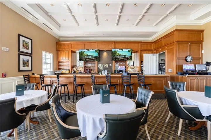 Lounge area featuring a coffered ceiling, wood-paneled bar with integrated televisions, patterned carpet flooring, and multiple round tables with upholstered seating