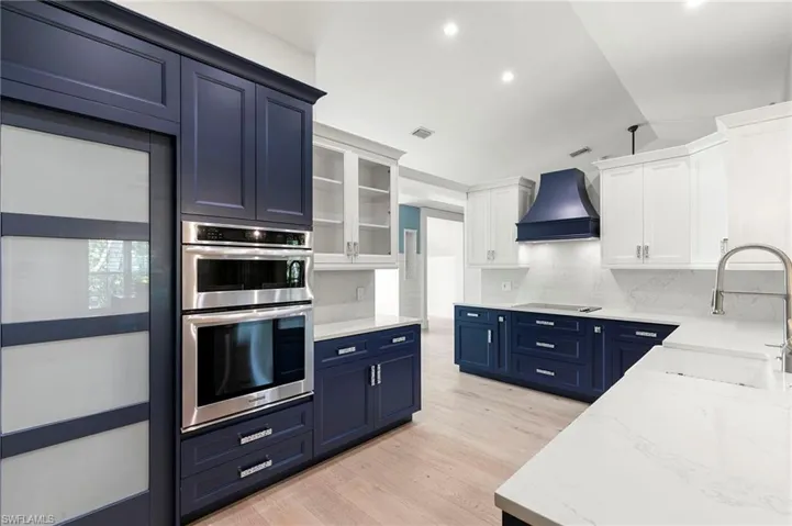 Kitchen with blue cabinets, stainless steel double oven, light stone counters, light wood-style flooring, and custom range hood